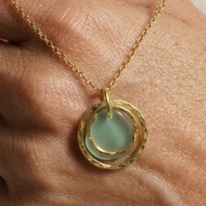 May include: A gold-colored necklace featuring a pendant with two concentric rings and a light green center. The necklace is held in a hand, showcasing the delicate chain and pendant design. The pendant has a hammered texture.