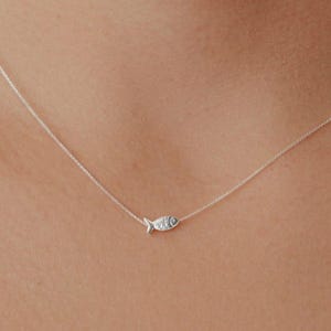 May include: A delicate silver necklace featuring a small fish pendant. The pendant is detailed with etched lines and a small eye. The necklace is shown against a skin tone background.
