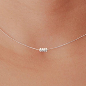 May include: A delicate silver necklace featuring three small, round beads centered on a fine chain. The necklace is displayed against a neutral skin tone, highlighting the minimalist design and subtle elegance of the jewelry.