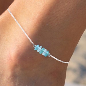 May include: A silver anklet with a light blue gemstone charm. The anklet is on a person's ankle.