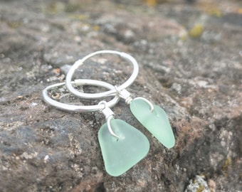 May include: A pair of silver hoop earrings with light green sea glass charms. The earrings are photographed on a rocky surface.