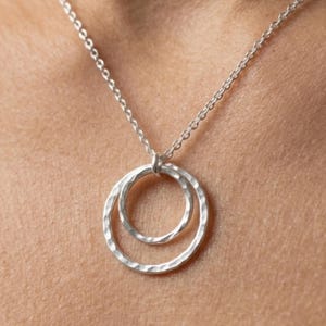 May include: A silver necklace featuring two textured, overlapping circles suspended from a delicate chain. The circles have a hammered finish, creating a subtle, reflective surface. The necklace is designed to be worn around the neck.