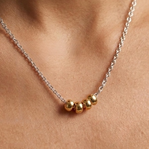 May include: A silver chain necklace with five gold-colored beads. The necklace is worn around the neck and the beads are evenly spaced. The chain is delicate and the beads are small and round.
