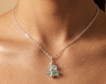 May include: A silver chain necklace featuring a pendant composed of two small, frosted light blue sea glass pieces separated and capped by tiny silver beads, creating a vertical stack design that rests against the collarbone area.