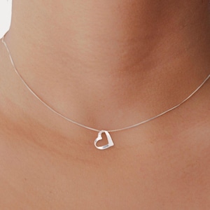 May include: A delicate silver necklace featuring a small, open heart pendant. The heart is a simple, minimalist design, suspended from a fine chain. The necklace is shown against a skin tone background, highlighting the jewelry's subtle elegance.