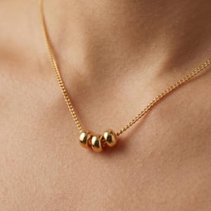 May include: A gold necklace with three small, spherical beads. The beads are strung on a fine chain. The necklace is shown against a skin-toned background, emphasising the jewellery's understated elegance.