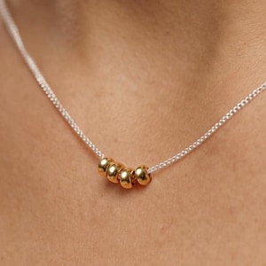 May include: A delicate silver necklace featuring five small, gold-colored beads. The beads are evenly spaced along the fine chain, creating a minimalist and elegant design. The necklace is shown against a skin tone background.