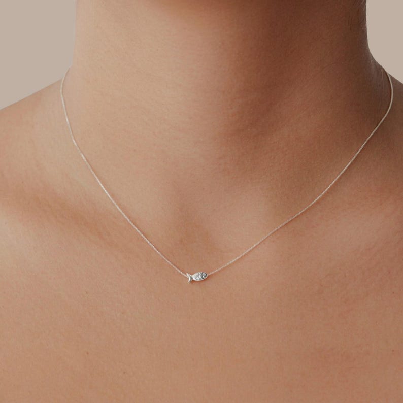 May include: A delicate silver necklace with a small fish pendant. The pendant has etched details and a tiny eye. The necklace is displayed on a person's neck.