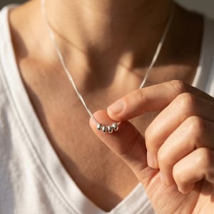 Może przedstawiać: Delikatny naszyjnik na srebrnym łańcuszku z czterema małymi, gładkimi, okrągłymi srebrnymi koralikami nawleczonymi na środku. Naszyjnik posiada cienki, polerowany łańcuszek z metalowych ogniw, kt&oacute;ry układa się na szyi, prezentując prosty design.