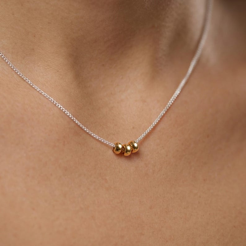 May include: A delicate necklace featuring a silver chain with three small, gold-colored beads. The beads are clustered together, creating a minimalist and elegant design. The necklace is shown against a skin tone background.