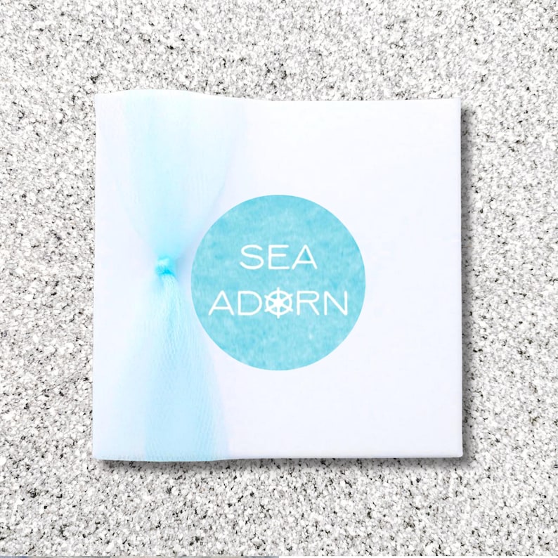 May include: A white square package with a light blue tulle bow. A light blue circle with the words "SEA ADORN" and a ship's wheel design is centered on the package.