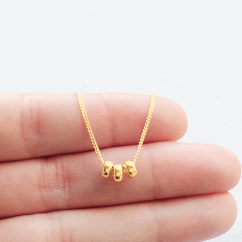 May include: A delicate gold necklace featuring three small, rounded beads. The beads are evenly spaced along a fine chain, creating a minimalist and elegant design. The necklace is shown against a neutral background, highlighting its simple beauty.