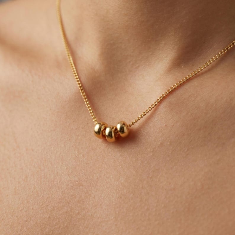 May include: A gold-colored necklace featuring three small, round beads. The necklace has a delicate chain and is worn around the neck. The beads are evenly spaced and have a polished finish, reflecting light.