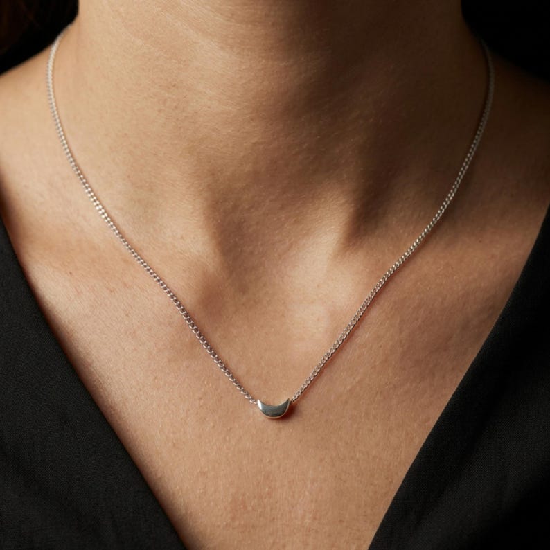 May include: A silver necklace with a small crescent moon pendant. The fine chain is displayed against a dark backdrop. The necklace is a simple, elegant piece of jewellery.