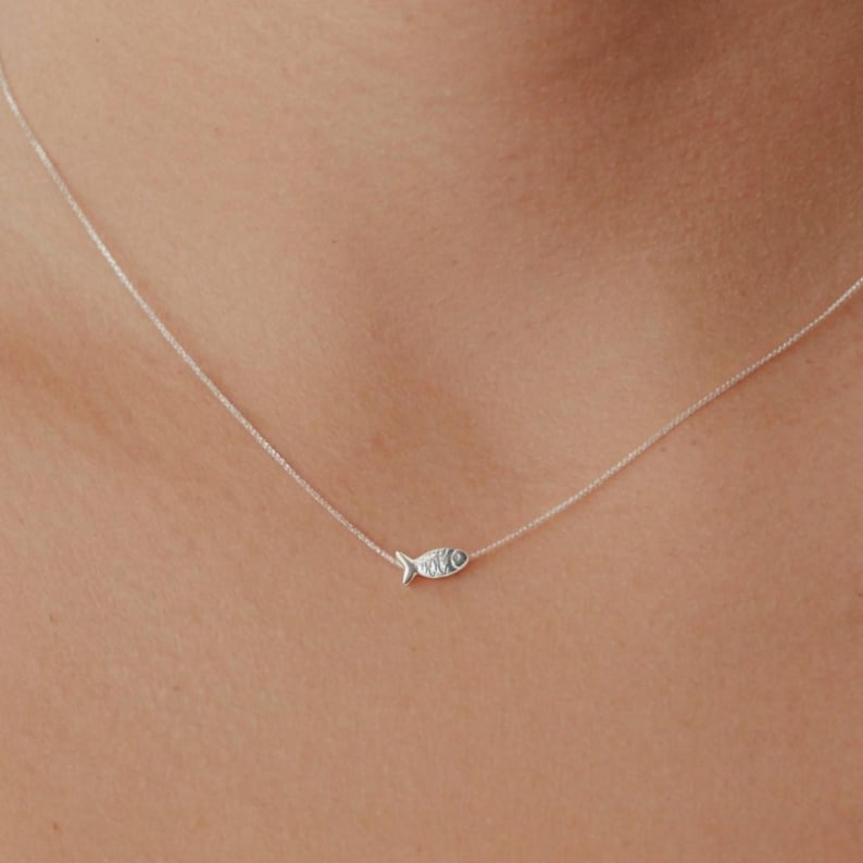May include: A delicate silver necklace featuring a small fish pendant. The pendant is detailed with fins and an eye, suspended on a fine chain. The necklace is shown against a skin tone background.