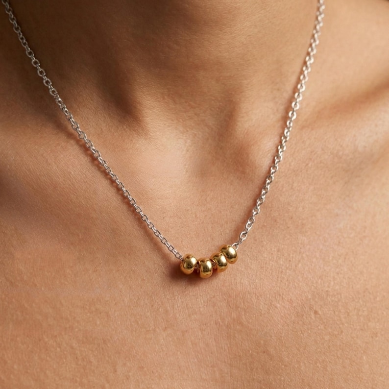 May include: A silver chain necklace with five gold-colored beads. The necklace is worn around the neck and the beads are evenly spaced. The chain is delicate and the beads are small and round.