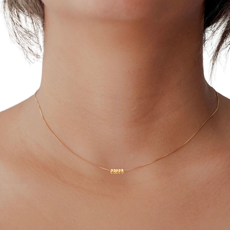 May include: A delicate gold necklace featuring a thin chain and a cluster of five small, round beads. The necklace is worn around the neck, showcasing its minimalist design and subtle elegance. The gold color provides a warm, classic look.
