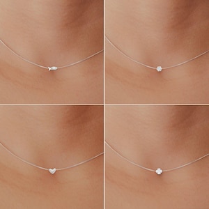 May include: Four delicate necklaces with different pendants: a fish, a small flower, a heart, and a four-leaf clover. Each pendant is suspended on a thin, nearly invisible chain, creating a floating effect. The pendants are silver-toned and small in size.