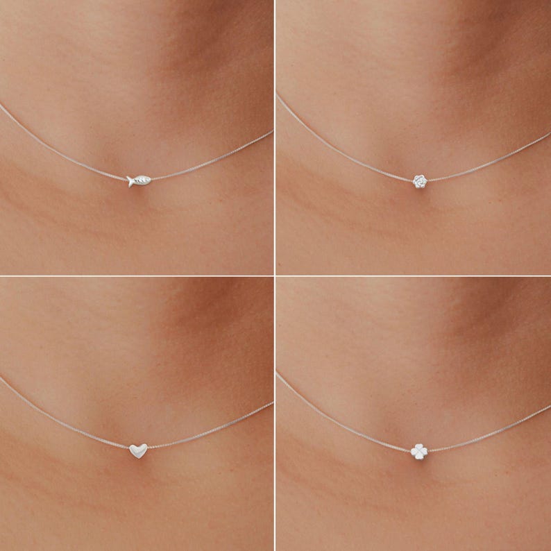 May include: Four delicate necklaces with different pendants: a fish, a small flower, a heart, and a four-leaf clover. Each pendant is suspended on a thin, nearly invisible chain, creating a floating effect. The pendants are silver-toned and small in size.