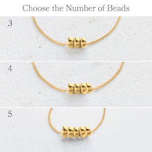 May include: Gold-colored necklaces with varying numbers of beads: three, four, and five. The text "Choose the Number of Beads" is at the top. The necklaces are displayed on a light-colored surface.