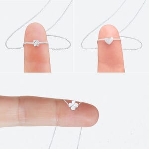 May include: Three delicate silver necklaces, each with a different pendant: a rose, a heart, and a four-leaf clover. The pendants are small and attached to a fine silver chain. The necklaces are displayed on a finger.