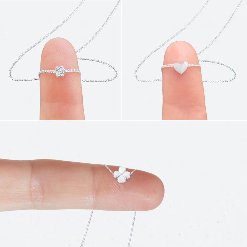 May include: Three delicate silver necklaces, each with a different pendant: a rose, a heart, and a four-leaf clover. The pendants are small and attached to a fine silver chain. The necklaces are displayed on a finger.