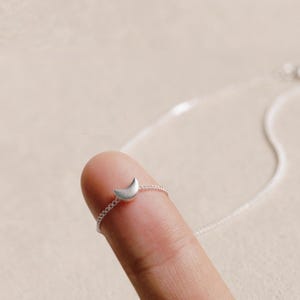 May include: A silver necklace with a crescent moon pendant. The small, curved moon is attached to a fine chain. The necklace is displayed on a finger, showing its size and design.