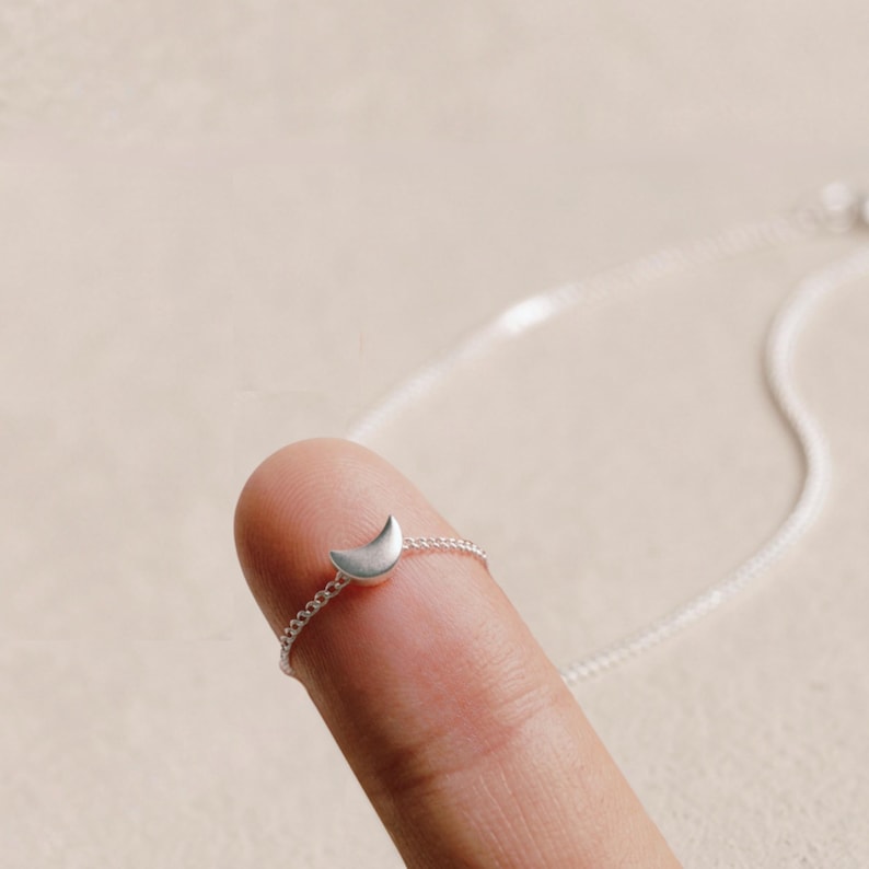 May include: A silver necklace with a crescent moon pendant. The small, curved moon is attached to a fine chain. The necklace is displayed on a finger, showing its size and design.