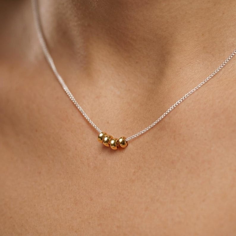 May include: A delicate silver necklace featuring five small, gold-colored beads. The beads are evenly spaced along the fine chain, creating a minimalist and elegant design. The necklace is shown against a skin tone background.