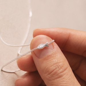 May include: A silver necklace with a tiny fish pendant. The fish charm has etched details and is suspended on a fine chain. The necklace is held between fingers, highlighting the jewellery's design and craftsmanship. The chain is a delicate curb style.