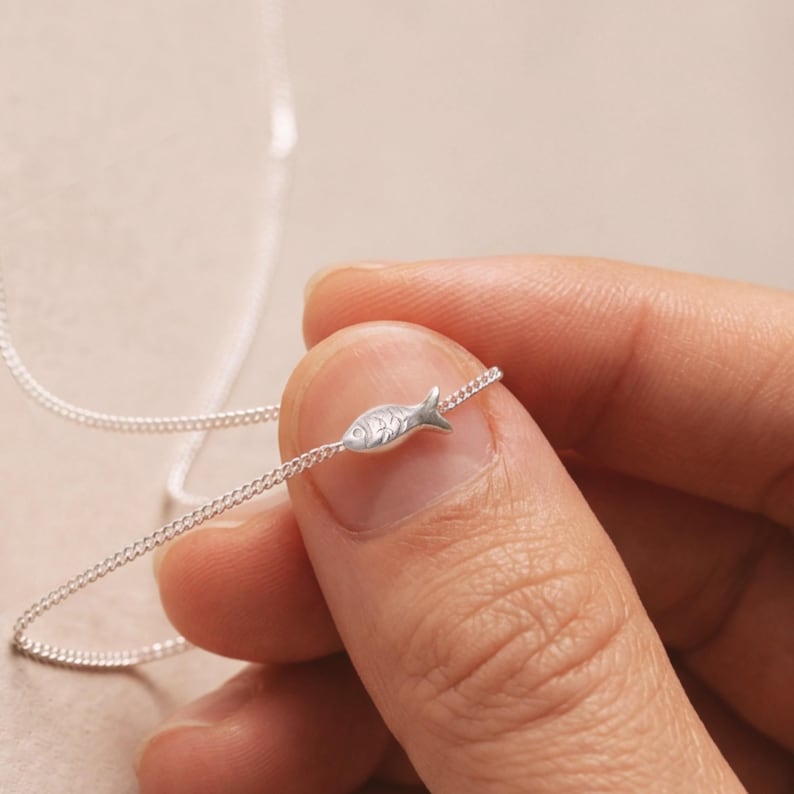 May include: A silver necklace with a tiny fish pendant. The fish charm has etched details and is suspended on a fine chain. The necklace is held between fingers, highlighting the jewellery's design and craftsmanship. The chain is a delicate curb style.