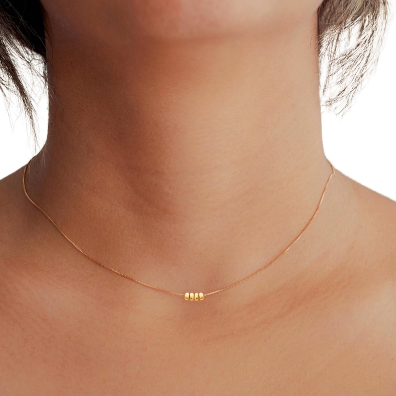 May include: A delicate gold necklace featuring three small, round beads. The necklace is displayed against a skin tone background, highlighting the simple elegance of the jewelry. The chain is thin and subtle, creating a minimalist aesthetic.