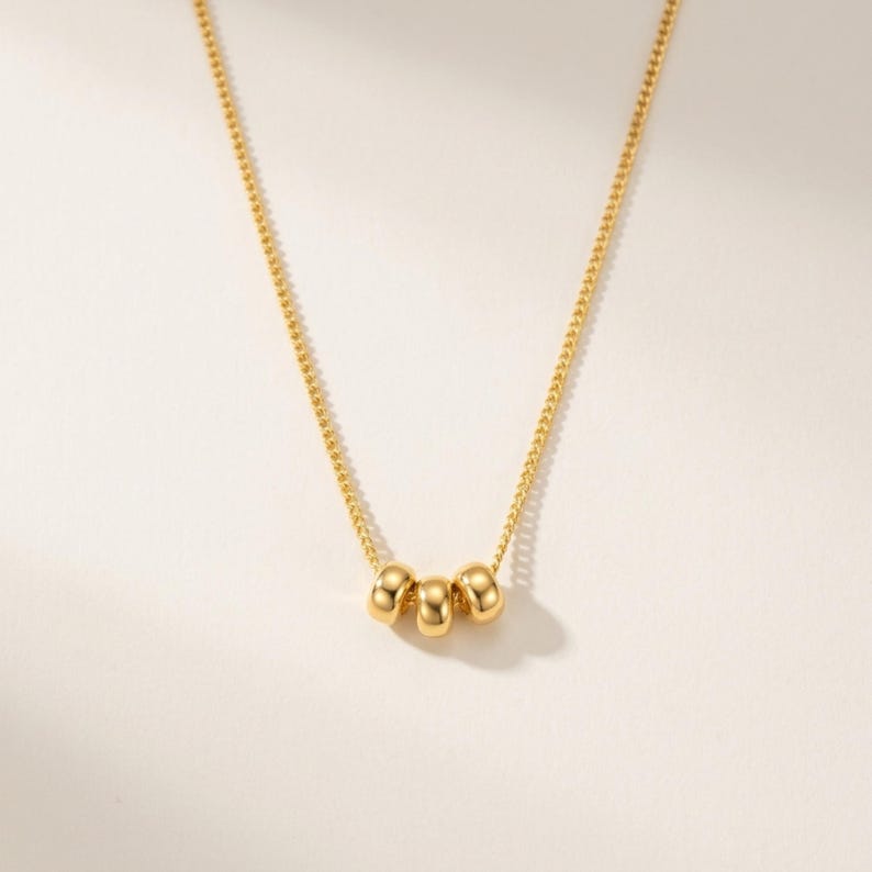 May include: A delicate gold necklace featuring three small, round beads. The beads are centered on a fine chain, creating a minimalist and elegant design. The necklace is displayed against a soft, neutral background, highlighting its simple beauty.