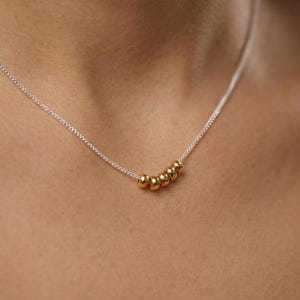 May include: A delicate necklace featuring a silver chain with six small, gold-colored beads. The necklace is worn around the neck, showcasing the minimalist design and the contrast between the silver chain and the gold beads.