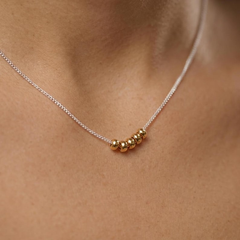 May include: A delicate necklace featuring a silver chain with six small, gold-colored beads. The necklace is worn around the neck, showcasing the minimalist design and the contrast between the silver chain and the gold beads.