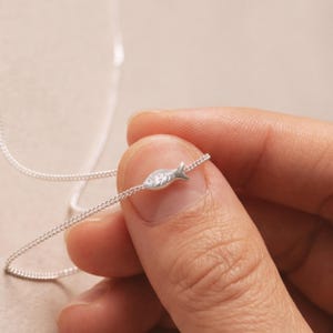 May include: A delicate silver necklace featuring a small fish pendant. The fish charm is detailed with etched scales and fins, suspended on a fine chain. The necklace is held between fingers, showcasing its dainty design and craftsmanship.