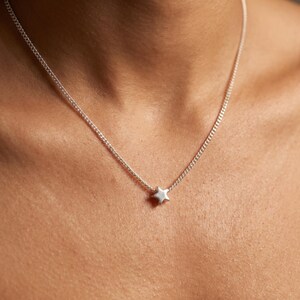 May include: A silver necklace with a small star pendant. The chain is fine and sits around the neck. The star is in the centre and appears to be made of a shiny metal.