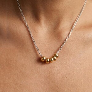 May include: A silver chain necklace with six gold-colored beads. The beads are evenly spaced along the bottom of the necklace. The necklace is worn around the neck.