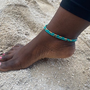 May include: A turquoise and gold beaded ankle bracelet. The bracelet is made of small beads and is worn on a person's ankle.