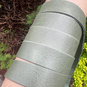May include: Close-up of a roll of olive green leather strips. The leather has a textured surface and is rolled around a cardboard tube. The edges of the leather are dark. The background includes green foliage.