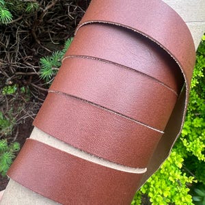 May include: Close-up of a rolled brown leather strip. The leather is a rich, reddish-brown color with a textured surface. The edges of the leather are cleanly cut, revealing a lighter tan interior. The leather is wrapped around a cardboard tube.