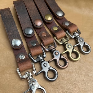 May include: Five leather keychains with different coloured metal clasps and rivets. The keychains are brown and have a distressed look.