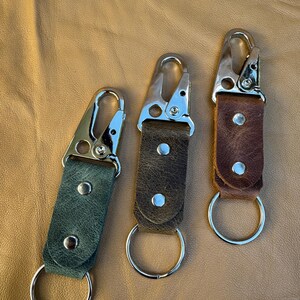 May include: Three keychains featuring textured leather straps in dark green, dark brown, and medium brown. Each strap is secured with two silver-toned metal rivets and attached to a silver-toned metal lobster clasp and a circular split ring.