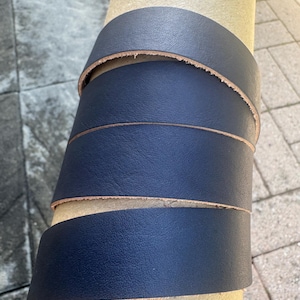 May include: A long, continuous strip of dark navy blue leather wrapped spirally around a cylindrical cardboard tube. The leather strap features a smooth, matte-textured surface on the exterior and a natural, light tan raw edge along the sides.