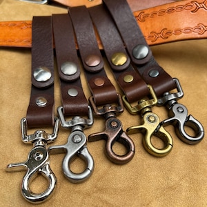 May include: Five leather keychains with different coloured metal clasps. The keychains are brown leather and have a metal loop at the top. The clasps are silver, copper, gold, and brass.