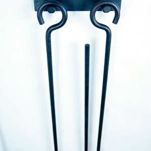 May include: Black metal fireplace tool holder with two hooks and a ring. The holder is mounted on a white wall.