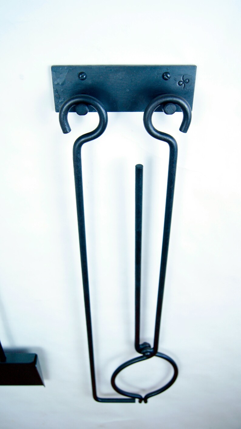 May include: Black metal fireplace tool holder with two hooks and a ring. The holder is mounted on a white wall.