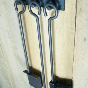 May include: A set of three black metal fireplace tools hanging on a wooden wall. The tools include a poker, a shovel, and a brush.