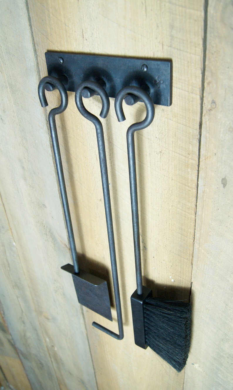 May include: A set of three black metal fireplace tools hanging on a wooden wall. The tools include a poker, a shovel, and a brush.