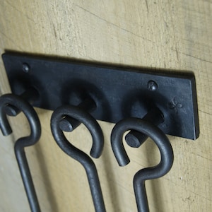 May include: Black metal fireplace tool holder with three hooks. The holder is mounted on a wooden wall.